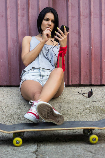 Hipster girl sitting on her skateboard and using smartphone - Foto, Bild