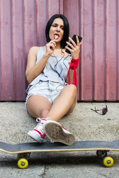 Hipster girl sitting on her skateboard and using smartphone - Фото, изображение