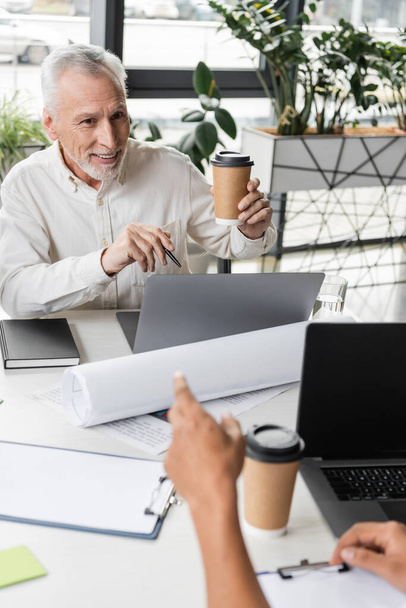 Positive middle aged businessman holding coffee to go near laptops and african american colleague in office  - Foto, Imagem
