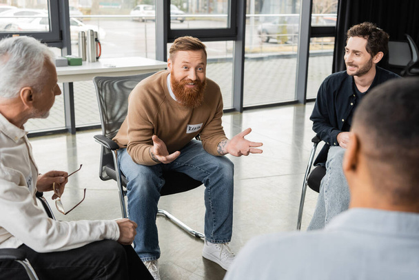 Hombre alegre con adicción al alcohol hablando con grupo interracial en centro de rehabilitación  - Foto, Imagen