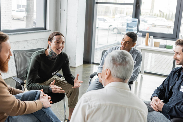 Homme tatoué joyeux avec dépendance à l'alcool parlant à un groupe interracial pendant la thérapie dans un centre de réadaptation  - Photo, image