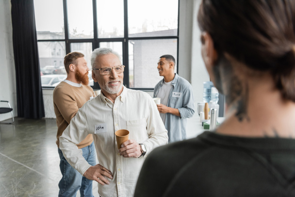 Sorrindo homem maduro com copo de papel e nome adesivo passar tempo durante alcoólatras reunião no centro de reabilitação  - Foto, Imagem