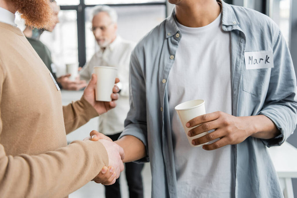 Hommes interraciaux avec des tasses en papier serrant la main lors d'une réunion d'alcooliques dans un centre de réadaptation  - Photo, image