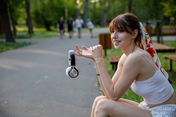 A happy girl sits on a park bench. She is holding wireless headphones in front of her. There are lots of trees and greenery around. In the distance you can see fuzzy figures of people walking. - Photo, Image
