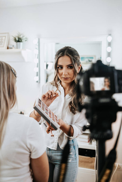 Happy young woman streaming a beauty makeup vlog from home or workshop. Beautiful online content creator cosmetician applying makeup and explaining some work tools. Vlogging and online channel work.  - Fotoğraf, Görsel