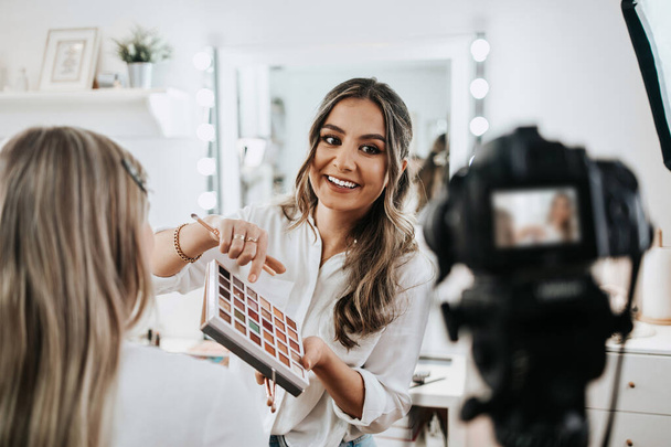 Happy young woman streaming a beauty makeup vlog from home or workshop. Beautiful online content creator cosmetician applying makeup and explaining some work tools. Vlogging and online channel work.  - 写真・画像