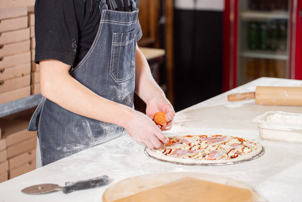 Closeup hand of chef baker in uniform blue apron cutting pizza at kitchen. High quality photo - Zdjęcie, obraz