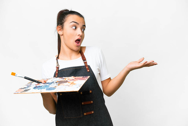Young artist woman holding a palette over isolated white background with surprise expression while looking side - Fotografie, Obrázek