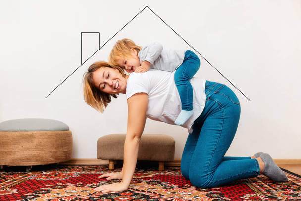 A happy mother and her little baby are playing in their new home. The symbol of the roof of the house is depicted above the family. The concept of mortgage and purchase of new housing. - Fotoğraf, Görsel