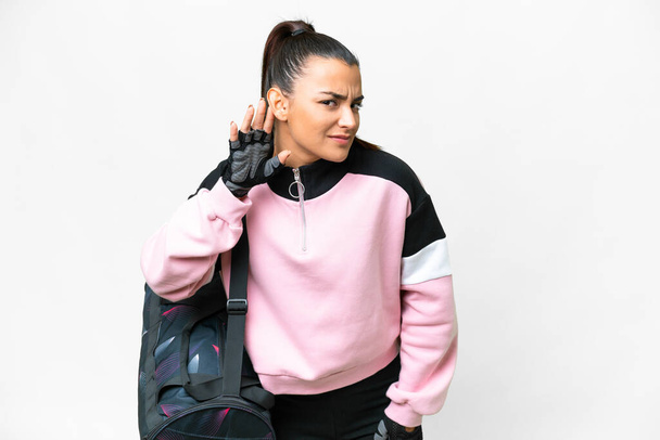 Young sport woman with sport bag over isolated white background listening to something by putting hand on the ear - Photo, Image