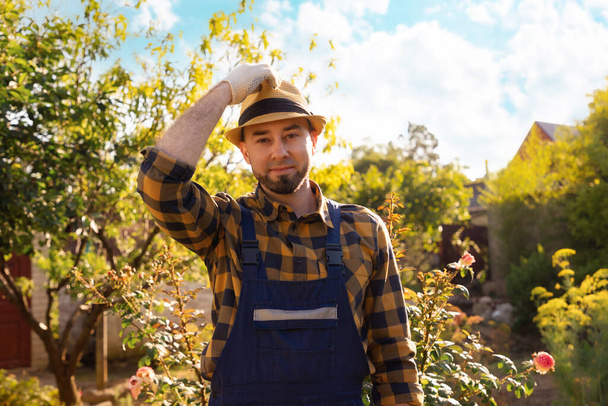 Portrait of a bearded young smiling gardener wearing a straw hat. In the background there is a backyard and a garden. The concept of gardening and horticulture. - Foto, Bild