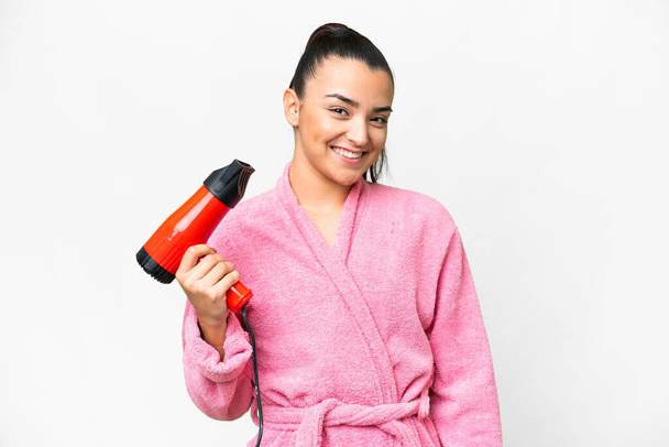 Young woman in a bathrobe holding a hairdryer over isolated white background smiling a lot - Foto, Imagen