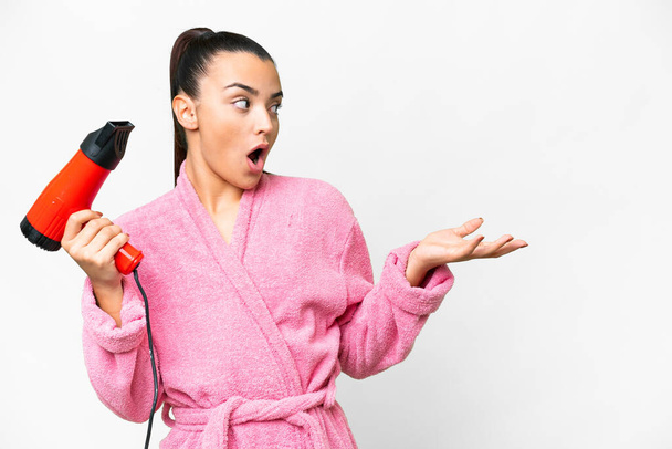 Young woman in a bathrobe holding a hairdryer over isolated white background with surprise facial expression - Foto, Imagem