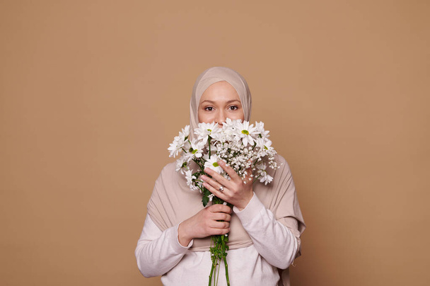 Charming Middle-Eastern Muslim woman with head covered in beige hijab, holding bouquet of white flowers covering half of her face, looking at camera with insightful gaze over isolated cream background - Photo, image