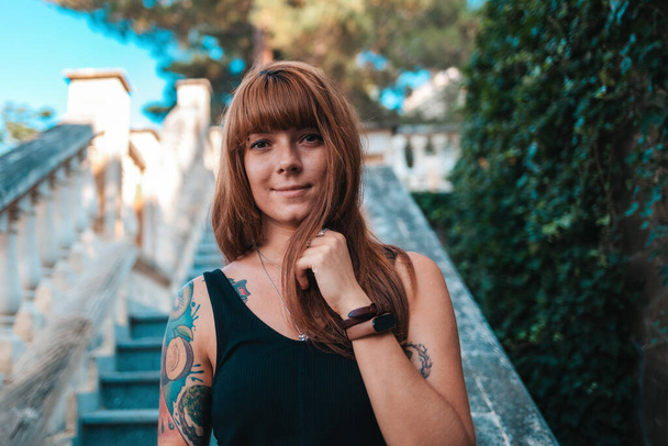 Retrato de una joven mujer bronceada sonriente con manga de tatuaje posando en los escalones de piedra. Concepto de psicología. - Foto, imagen