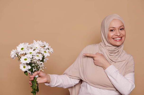 Приваблива елегантна арабська мусульманка в хіджабі, усміхаючись зубатою посмішкою, вказуючи на прекрасний букет білих хризантем і квіток гіпсофіли, дивлячись на камеру, ізольовану на бежевому тлі. - Фото, зображення