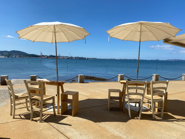 два деревянных винтажных стола со стульями и зонтиком ждут гостей в жаркое лето перед океаном - Фото, изображение