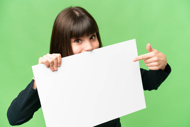 Little girl over isolated chroma key background holding an empty placard and hiding behind it - Photo, Image