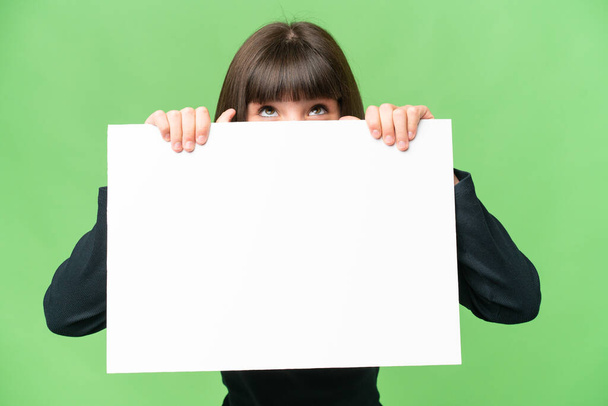 Menina sobre fundo chave croma isolado segurando um cartaz vazio e se escondendo atrás dele - Foto, Imagem