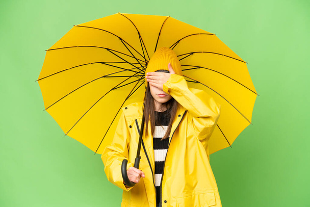 Little girl with rainproof coat and umbrella over isolated chroma key background covering eyes by hands. Do not want to see something - Foto, Imagem