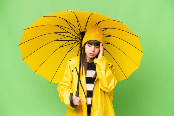 Маленькая девочка с непромокаемым плащом и зонтиком на изолированном фоне хрома ключ с головной болью - Фото, изображение