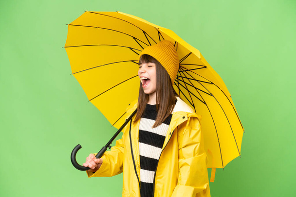 Niña con abrigo impermeable y paraguas sobre fondo de croma key aislado riendo en posición lateral - Foto, Imagen