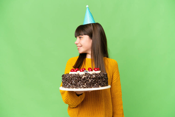 Little girl holding birthday cake over isolated chroma key background looking side - 写真・画像