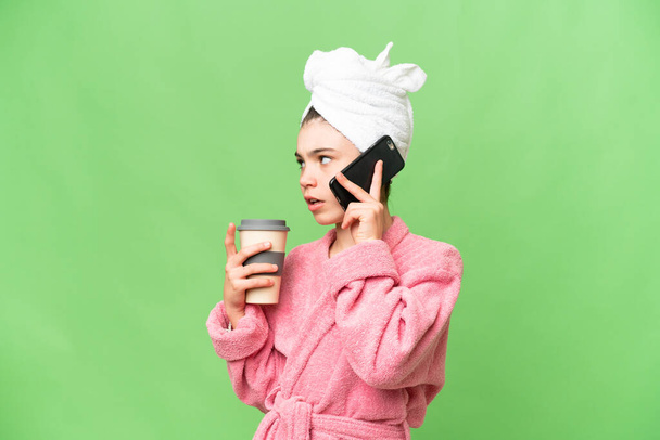 Petite fille en peignoir sur fond de chroma isolé tenant du café à emporter et un mobile - Photo, image