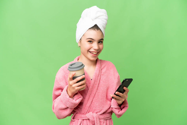 Klein meisje in een badjas over geïsoleerde chroma key achtergrond met koffie mee te nemen en een mobiele - Foto, afbeelding