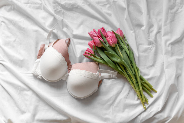 Weiße und rosa BHs, rosa Tulpen auf dem Bett. Frauen zarte Dessous, Unterwäsche. Von oben gesehen, aus nächster Nähe. Flachlage, Beauty-Blog oder Social Media Minimalkonzept. Geschenk zum Valentinstag, Frauentag - Foto, Bild