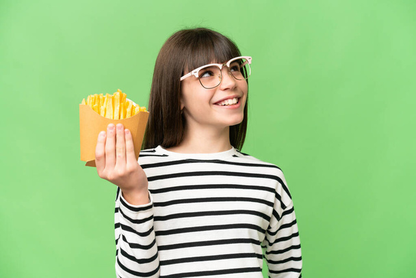 Μικρό κορίτσι που κρατάει τηγανητά πατατάκια πάνω από απομονωμένο φόντο chroma κλειδί κοιτάζοντας ψηλά, ενώ χαμογελά - Φωτογραφία, εικόνα