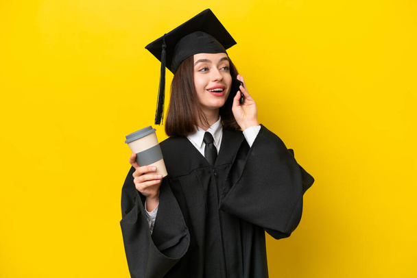 Jovem universitária graduada ucraniana mulher isolada no fundo amarelo segurando café para levar e um celular - Foto, Imagem