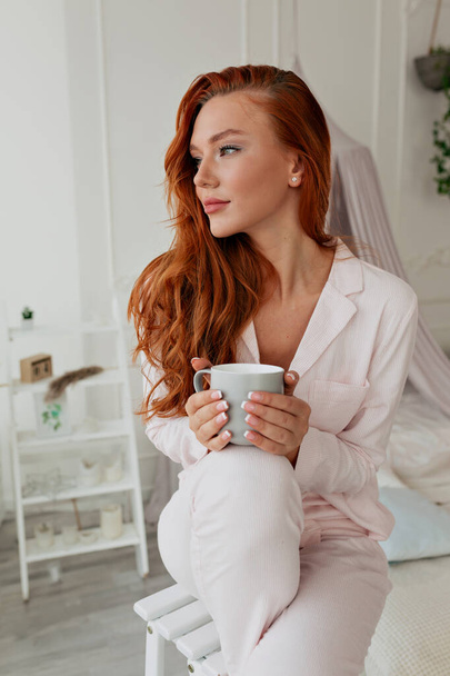 Spectaculaire schattige dame met lang rood haar draagt roze pyjama houdt 's ochtends koffie en kijkt naar het raam met een zachte glimlach in witte stijlvolle kamer. - Foto, afbeelding