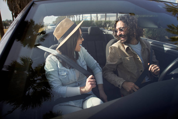 Νεαρό καυκάσιο ευτυχισμένο ζευγάρι που δένεται σε ένα κάμπριο αυτοκίνητο. Χαρούμενος γαμπρός με την όμορφη νύφη του στο όχημα. Χαμογελώντας άντρας και γυναίκα διασκεδάζουν μαζί σε ένα ταξίδι το Σαββατοκύριακο. Ζώνη ασφαλείας, ασφαλής οδήγηση. - Φωτογραφία, εικόνα