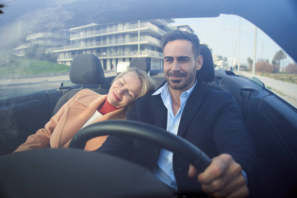 Μεσήλικες που οδηγούν αμάξι σε αστικό δρόμο. Ευτυχισμένη ξανθιά γυναίκα που στηρίζεται σε ελκυστικό άνδρα απολαμβάνοντας το αίσθημα της ελευθερίας των διακοπών. Άνθρωποι που οδηγούν ανέμελα στο τοπίο με την πόλη στο παρασκήνιο - Φωτογραφία, εικόνα