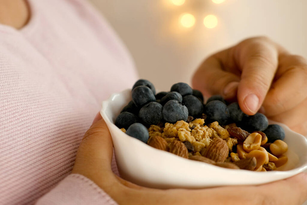 close-up female hand takes berries on blurred background with bokeh, healthy food white cup, oatmeal, blueberries, nuts, almonds, peanuts , raisins, dried fruits, raw food diet, vegetarian food - Foto, Imagen