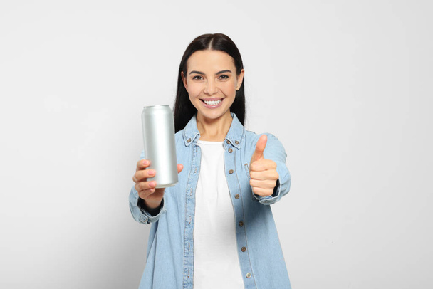 Beautiful happy woman holding beverage can and showing thumbs up on light grey background - Foto, afbeelding