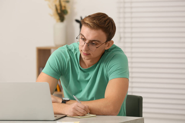 Young man with laptop writing in notebook at table indoors - Photo, image