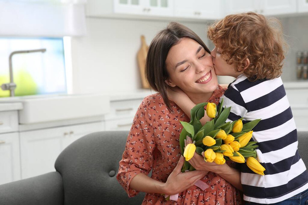 Little son kissing and congratulating his mom with Mother`s day at home, space for text. Woman holding bouquet of yellow tulips - Фото, изображение
