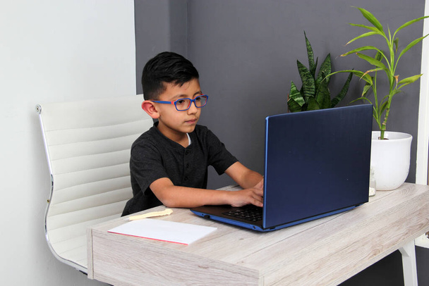 7-year-old Latino boy with glasses does home schooling takes online classes at home on a desk with a laptop, studies, is surprised and participates in class - Photo, Image