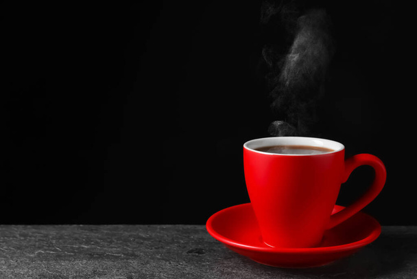Red cup with hot steaming coffee on grey table against black background, space for text - Photo, image