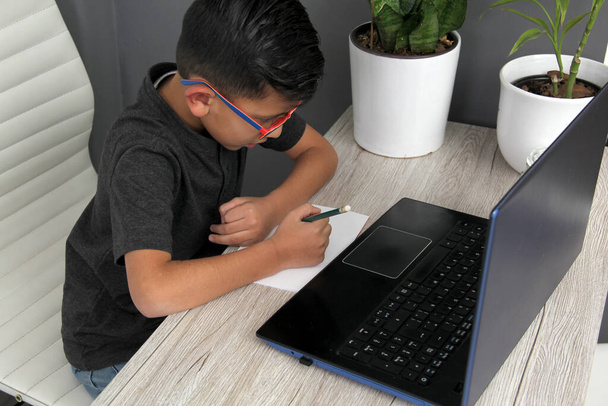 7-year-old Latino boy with glasses does home schooling takes online classes at home on a desk with a laptop, studies, is surprised and participates in class - Fotoğraf, Görsel
