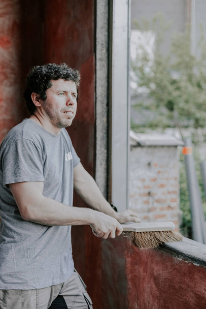 Young caucasian man holding an old broom and looking thoughtfully to the side sweeps a dirty window frame, closeup side view. The concept of cleaning and installing windows, construction work, house renovation. - Foto, immagini