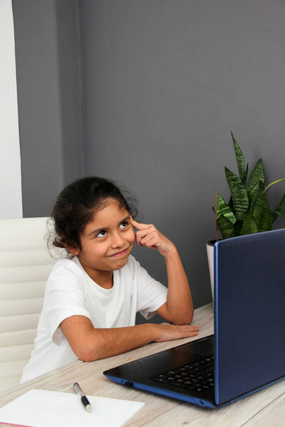 9-year-old Latino girl does home schooling takes online classes at home on a desk with a laptop, studies, is surprised and participates in class - Photo, Image