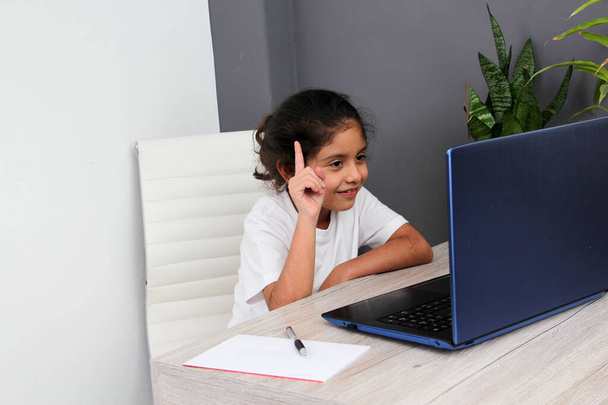 9-year-old Latino girl does home schooling takes online classes at home on a desk with a laptop, studies, is surprised and participates in class - Fotoğraf, Görsel