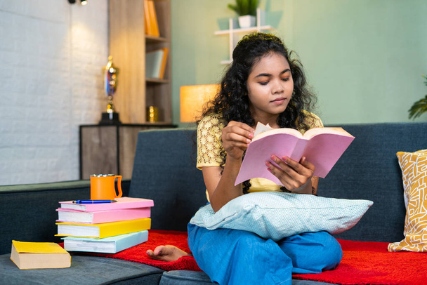 Vakava nuori tyttö lukeminen kirja tutkittavaksi istuessaan sohvalla kotona - käsite koulutus, valmistelu ja harrastukset. - Valokuva, kuva
