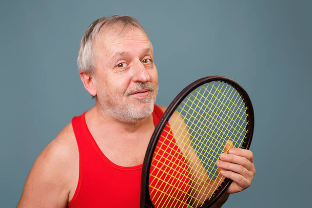Senior Men Enjoying Tennis A pensioner who is still active in sports, is captured in this image. The man is seen holding a tennis racket, dressed in a red tank top and is set against a blue background - Фото, изображение