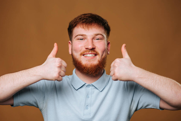 Un jeune rouquin donne un pouce vers le haut avec un sourire rayonnant dans un plan de studio, Adolescent mâle avec une expression joyeuse et un pouce vers le haut dans un portrait de studio - Photo, image