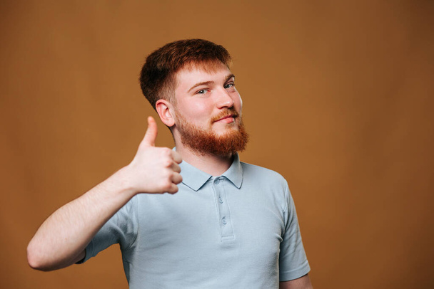 A teenage guy with a bright smile looks straight at the camera and gives a thumbs up in a studio setting. redhead teen shows his optimism and joy by flashing a thumbs up and smiling - Foto, Imagem