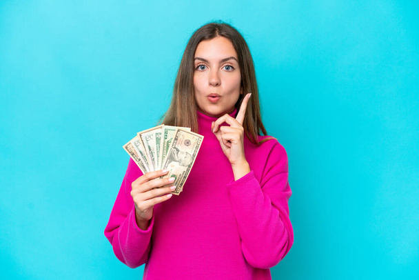 Nuori valkoihoinen nainen ottaa paljon rahaa eristetty sinisellä taustalla aikoo toteuttaa ratkaisun nostaessaan sormea ylös - Valokuva, kuva